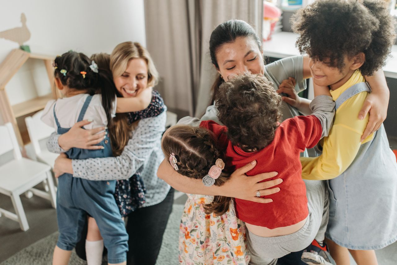 Children and women sharing a heartwarming group hug indoors, expressing love and diversity.